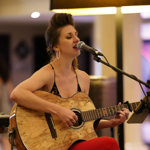 A woman is playing the guitar and singing into a microphone while seated in an indoor setting with a lamp in the background.