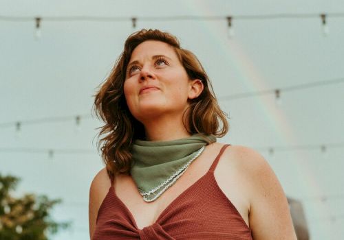 A person with a brown top and green scarf looks up, with string lights and a faint rainbow in the background under a cloudy sky.