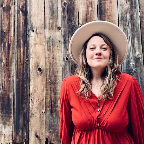 A person wearing a white hat and a red shirt stands in front of a rustic wooden wall, smiling at the camera.